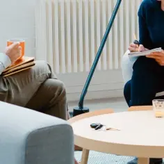 Dwie osoby siedzą w fotelach, jedna trzyma kubek, druga pisze w notatniku.