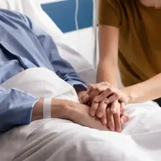 Opieka paliatywna co to? Dłoń bliskiej osoby dodaje otuchy pacjentowi w szpitalnym łóżku.