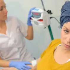 Kobieta w chustce na głowie przechodzi chemioterapię. Pielęgniarka przygotowuje aparaturę.