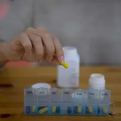 Dłoń umieszcza żółtą kapsułkę, być może zawierającą kwas bempediowy, w pojemniku na leki na cały tydzień.