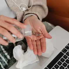 Dłonie wysypują kapsułki z butelki do dłoni. Obok leży laptop i chusteczki, sugerując chorobowe.