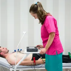 Pacjent leży na kozetce, pielęgniarka przygotowuje aparat do EKG. Badanie EKG jest szybkie i bezbolesne.