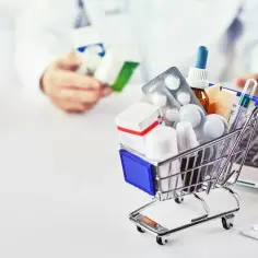 Mini wózek z lekami, termometr i opakowania tabletek. W tle farmaceuta przy komputerze, zapewne korzysta z systemu Haleon.