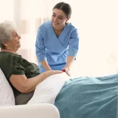 Uśmiechnięta opiekunka w niebieskim uniformie wspiera starszą pacjentkę w łóżku. To obraz troski, jaki oferuje hospicjum, zapewniając komfort i wsparcie.