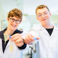 Dwóch młodych mężczyzn w białych fartuchach laboratoryjnych trzyma probówki.
