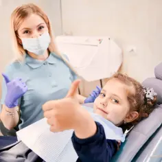 Uśmiechnięta dziewczynka w gabinecie dentystycznym, która otrzymała znieczulenie ogólne, pokazuje kciuk w górę. Obok niej stoi dentystka w masce i rękawiczkach, również z kciukiem w górę.
