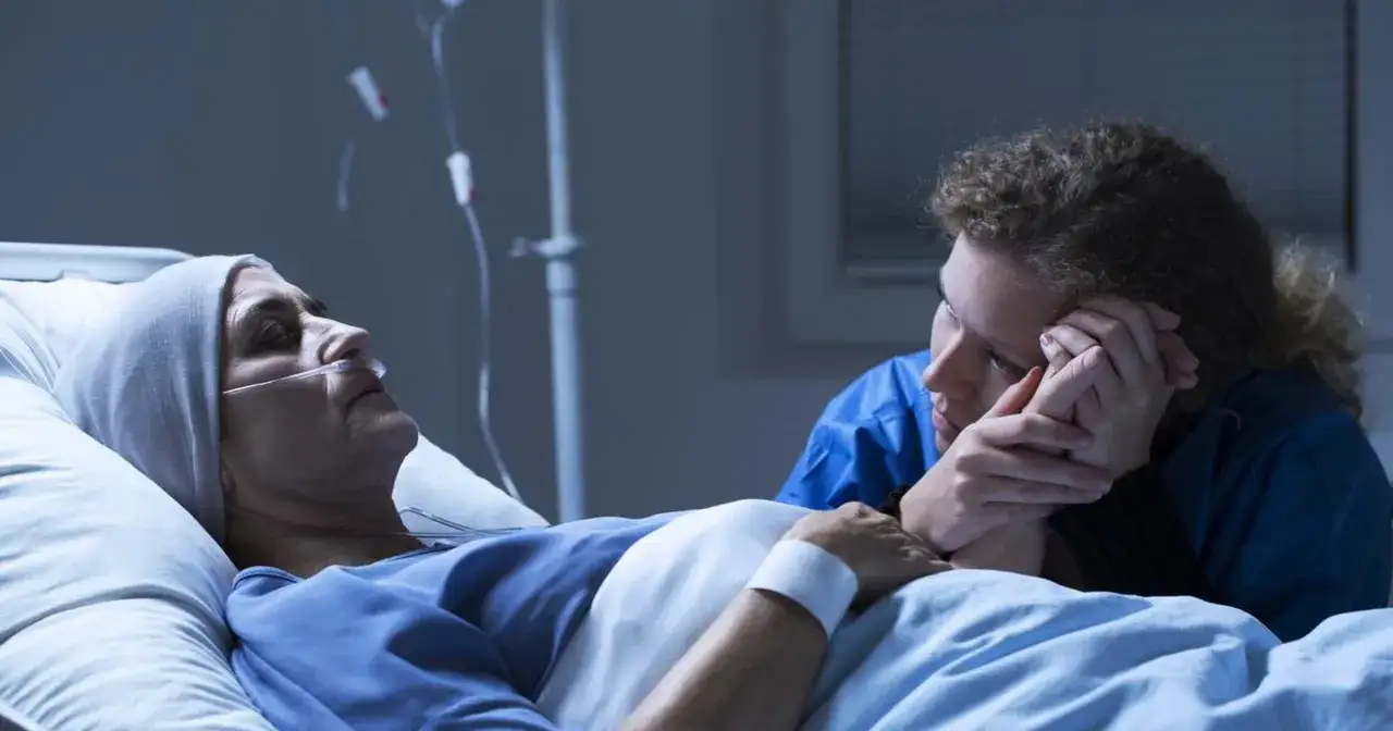 Na oddziale paliatywnym młoda kobieta trzyma za rękę umierającą matkę. W tle widać kroplówkę.