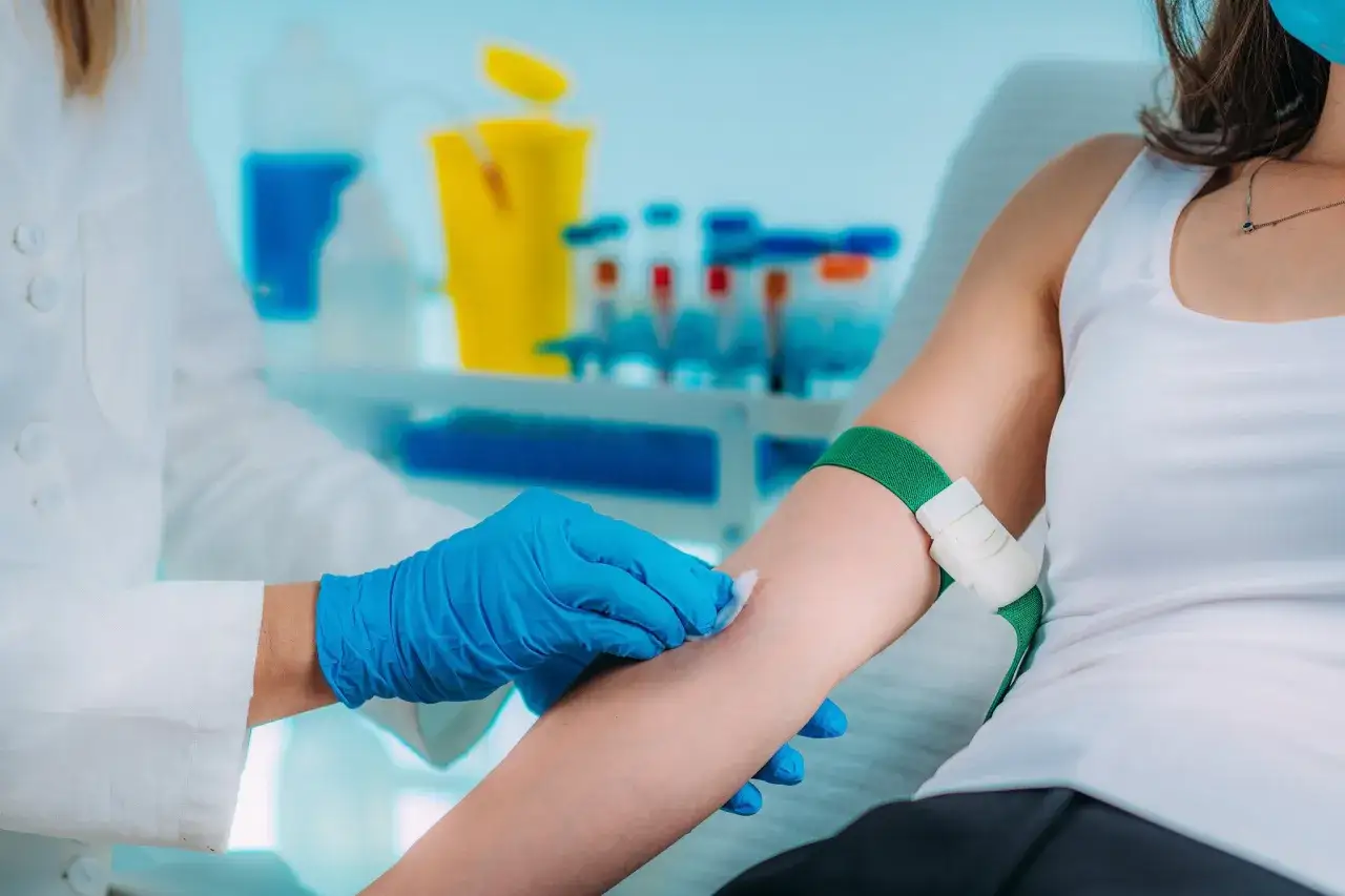 Person w rękawiczkach dezynfekuje ramię przed flebotomią. W tle probówki i sprzęt laboratoryjny.