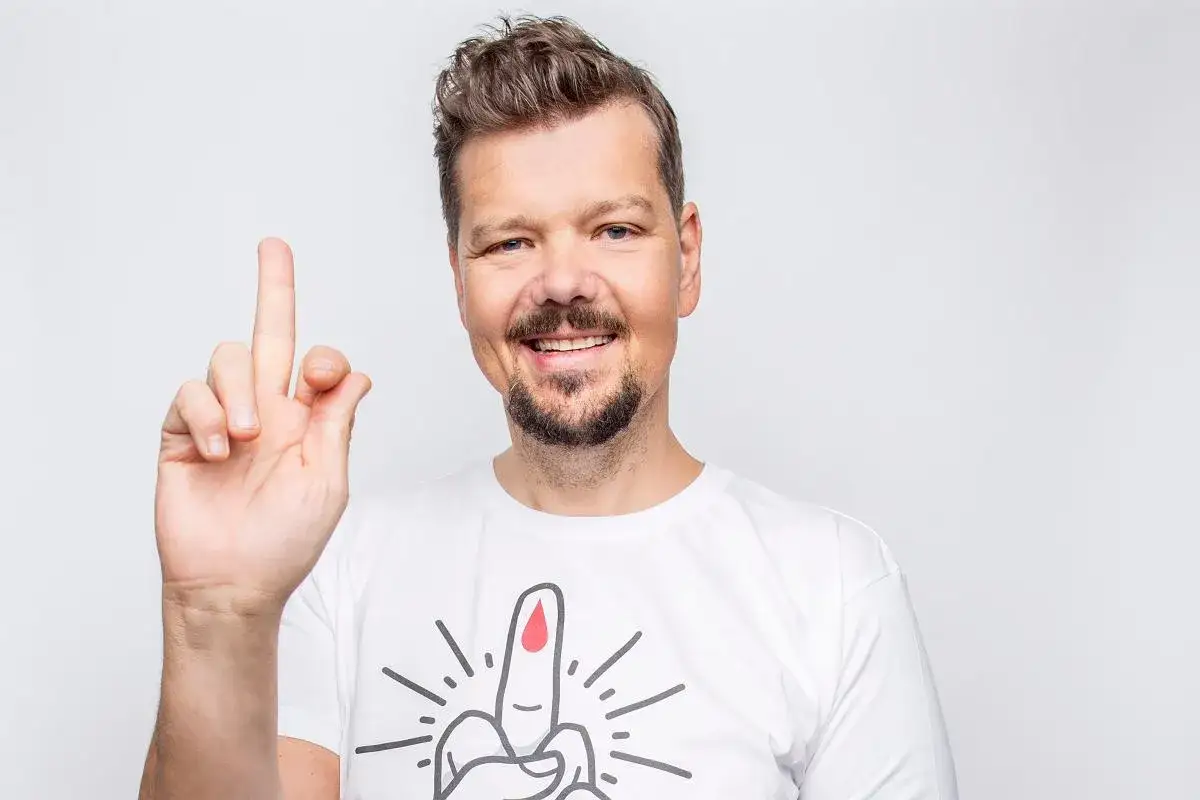 Uśmiechnięty mężczyzna z zarostem podnosi palec wskazujący. Na jego białej koszulce widnieje rysunek palca z kroplą krwi, symbolizujący nadzieję na przeszczep trzustki.
